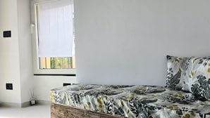 Ett rum med en säng, ett fönster med persienner, ett litet bord med ett ljus och en väggmonterad strömbrytare.