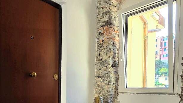 A room with a wooden door, a stone wall, a small wooden cabinet, and a window with a view of buildings outside. - The Pirate's Penthouse- Bluchalet  (Riomaggiore)