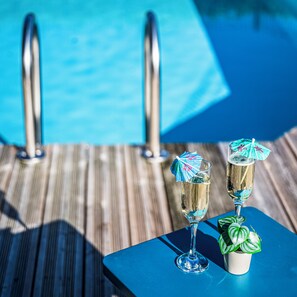 Ein Tisch am Pool mit zwei Sektgläsern und Sonnenschirmen, einer Topfpflanze sowie Liegestühlen.