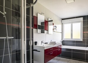Ein modernes Badezimmer mit einer Duschkabine aus Glas, einem weißen Waschbecken mit rotem Unterschrank und einem großen Spiegel darüber.