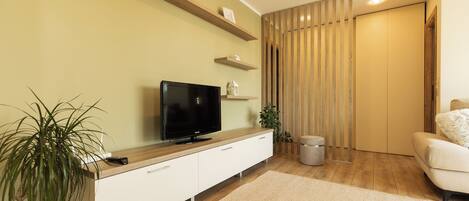 A modern living room with a flat-screen TV, wooden shelves, a potted plant, and a beige sofa.