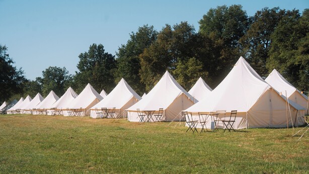 A row of white tents with tables and chairs, set in a grassy field with trees in the background.