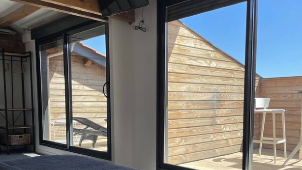 A bedroom with a large window, a bed with a quilt, a wooden ceiling, and a view of the backyard.