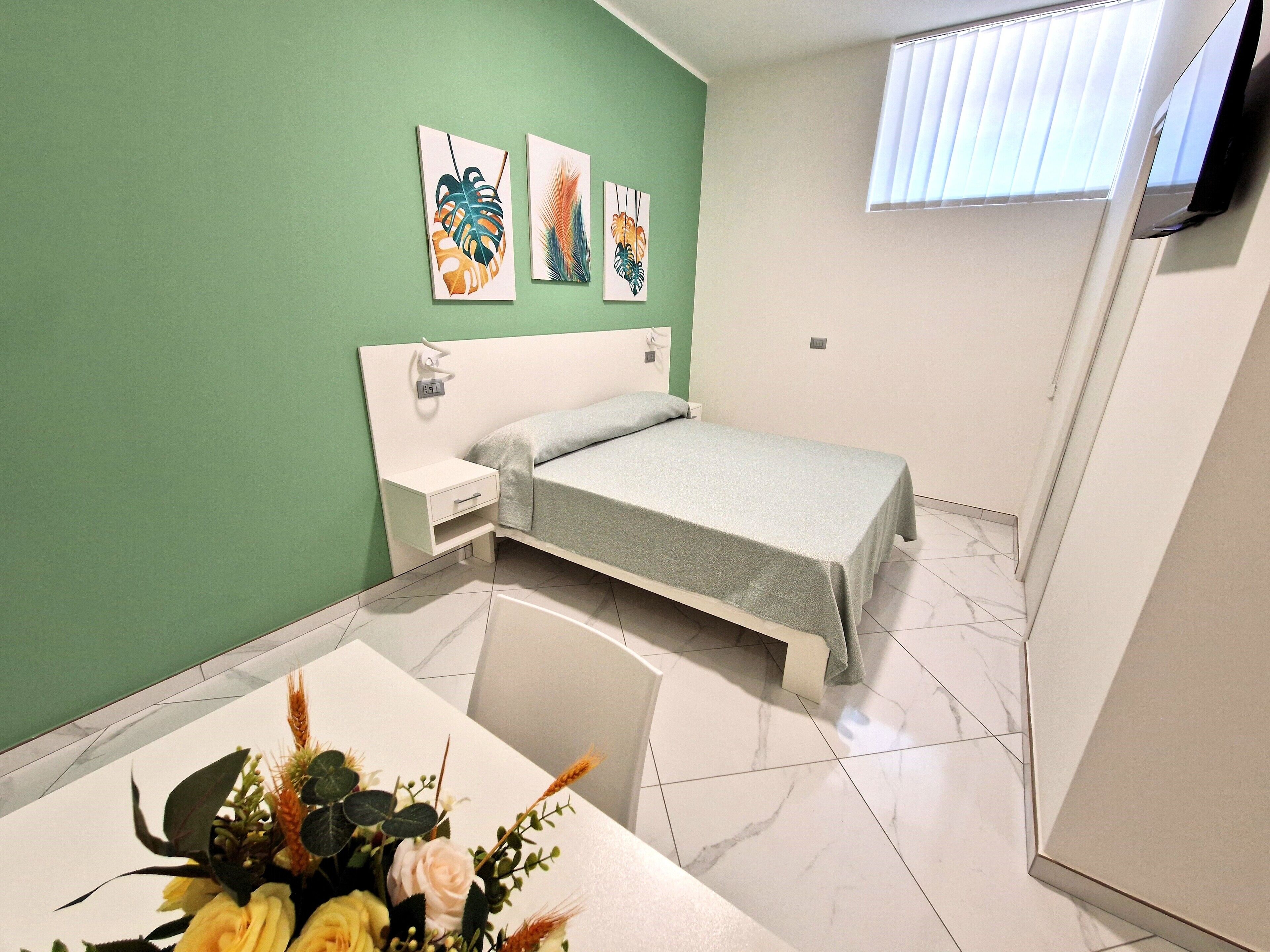 Een kleine, schone kamer met een bed, nachtkastje, een wandlamp en een raam met jaloezieën.