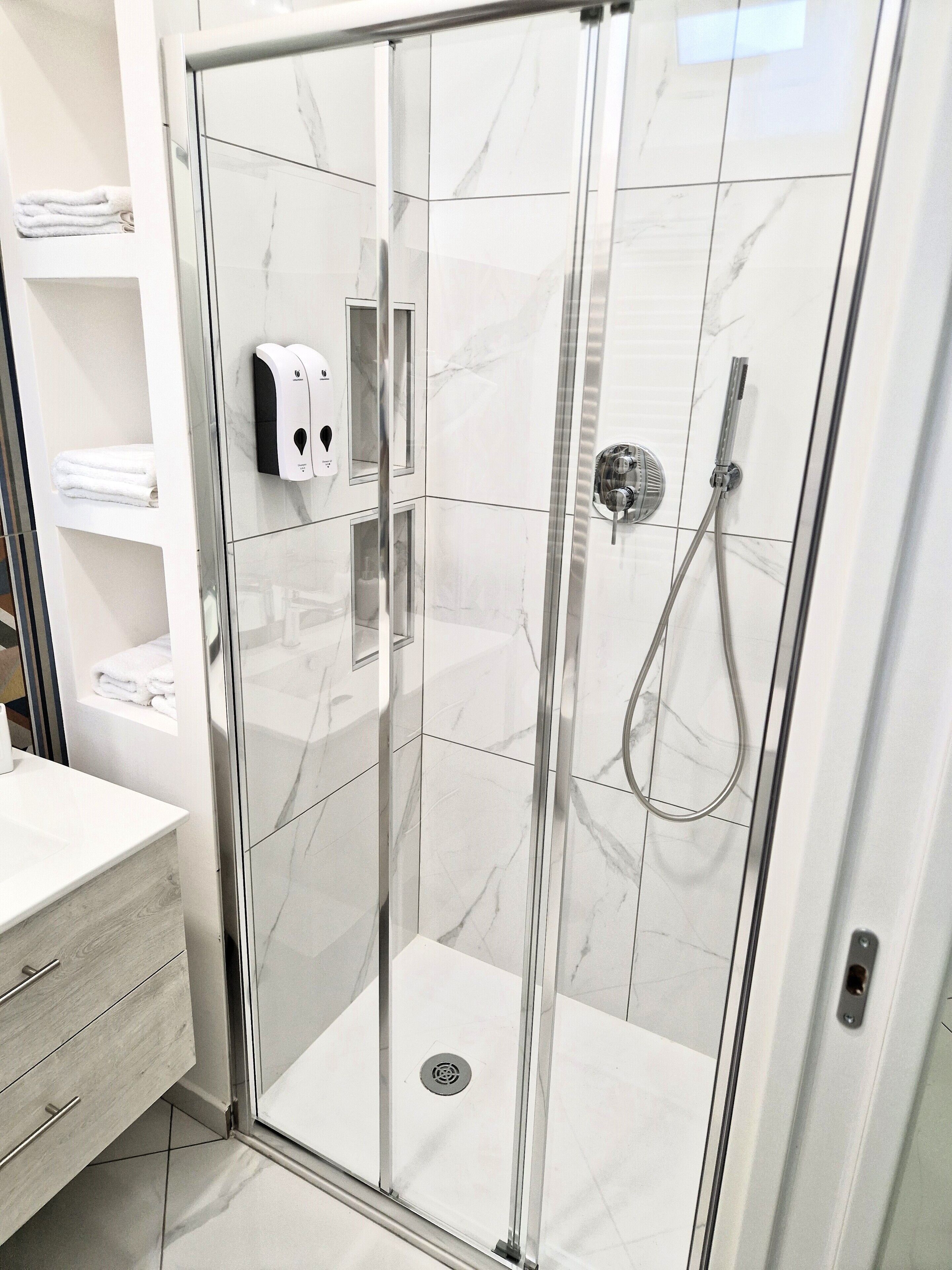 Une salle de bain moderne dotée d’une douche à parois de verre, d’un mur recouvert de carreaux de marbre blanc, d’un distributeur de serviettes intégré et d’un meuble-lavabo en bois avec des tiroirs.