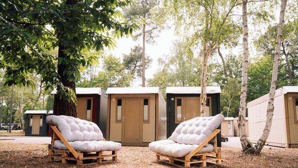 Zwei Holz-Loungesessel mit Kissen in einer bewaldeten Gegend, im Hintergrund tragbare Holzhütten.