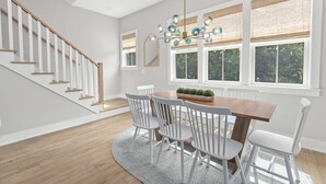 A dining area with a wooden table, white chairs, a chandelier, and large windows with blinds.