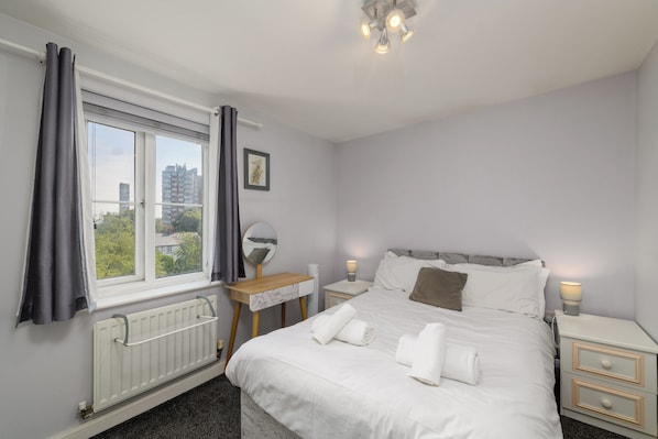 A bedroom with a bed, bedside tables, a desk, a radiator, and a window with curtains.