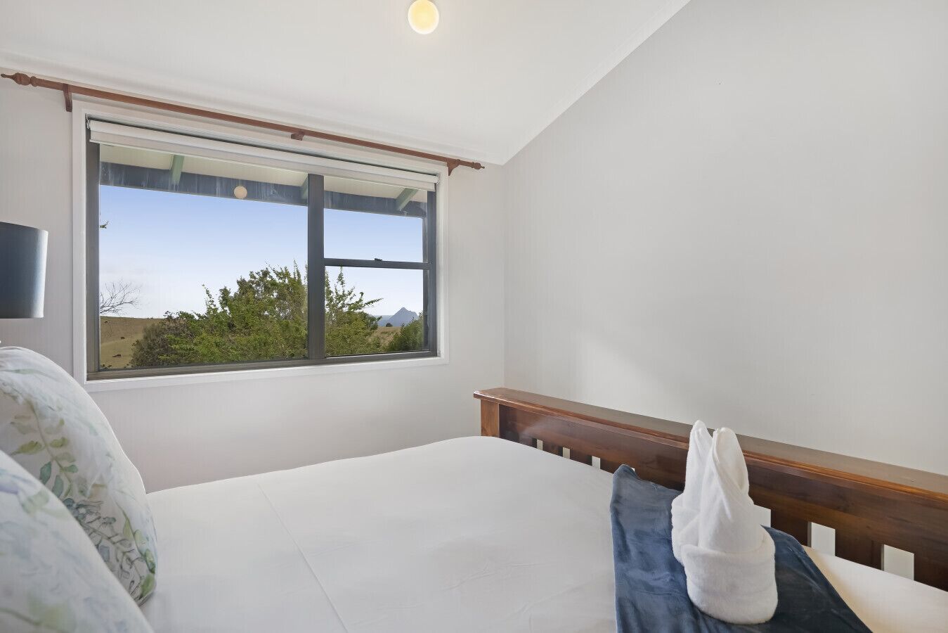 A bedroom with a large window, a bed, a wooden headboard, and a bedside table.