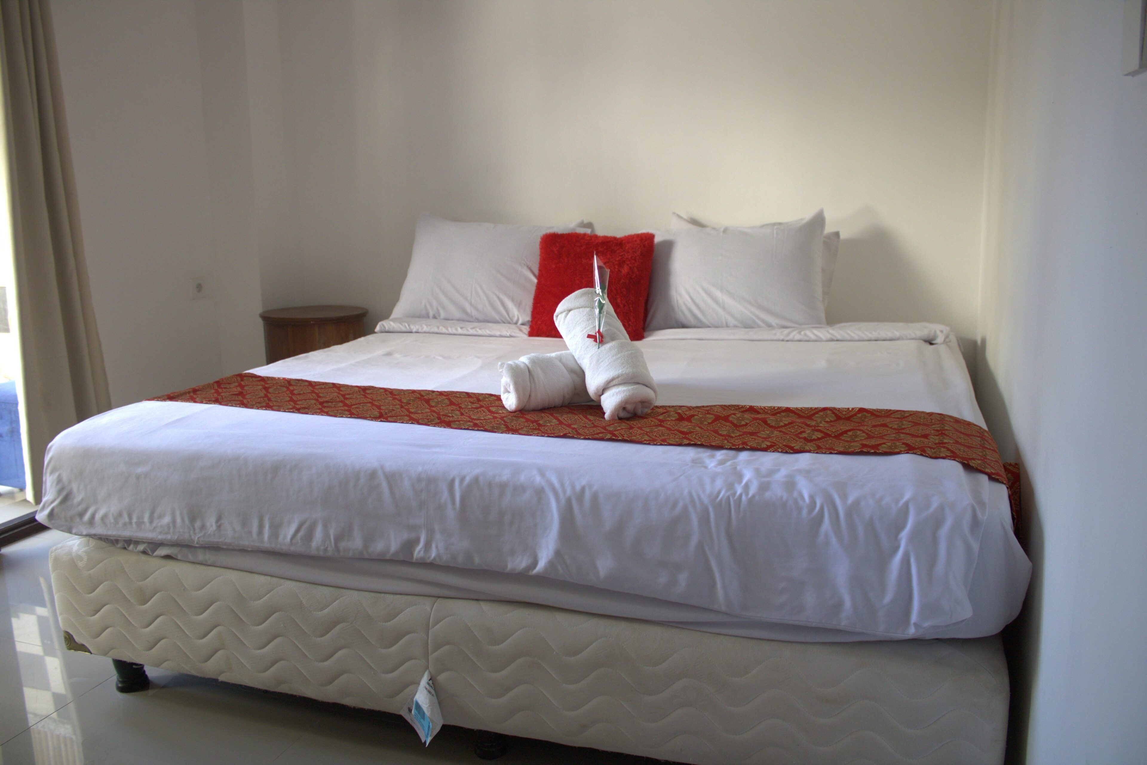 A neatly made bed with white bedding, a red throw pillow, and a white towel folded on top.