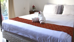 Una cama bien tendida con sábanas blancas y una colcha roja estampada, una mesita de noche con una flor, y un cuadro en la pared.