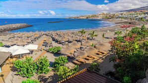 A beach resort with thatched umbrellas, sun loungers, and a view of the sea and town. - SeaShell Crown Luxury Suite (Costa Adeje)