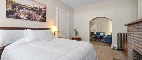 A hotel room with a large bed, a ceiling fan, a painting of a cityscape, a fireplace, and a living area with a sofa and a coffee table.