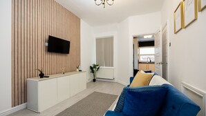 Ein modernes Wohnzimmer mit einem blauen Sofa, einem Flachbildfernseher an einer Wand aus Holzpaneelen, einem Heizkörper und Blick in die Küche durch eine offene Tür.