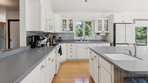 A modern kitchen with white cabinets, a granite countertop, and a large window providing a view of the outdoors. - Historic Charm Meets Modern Luxury in Kennebunkport|Walk to Shops & Restaurants! (Kennebunkport)