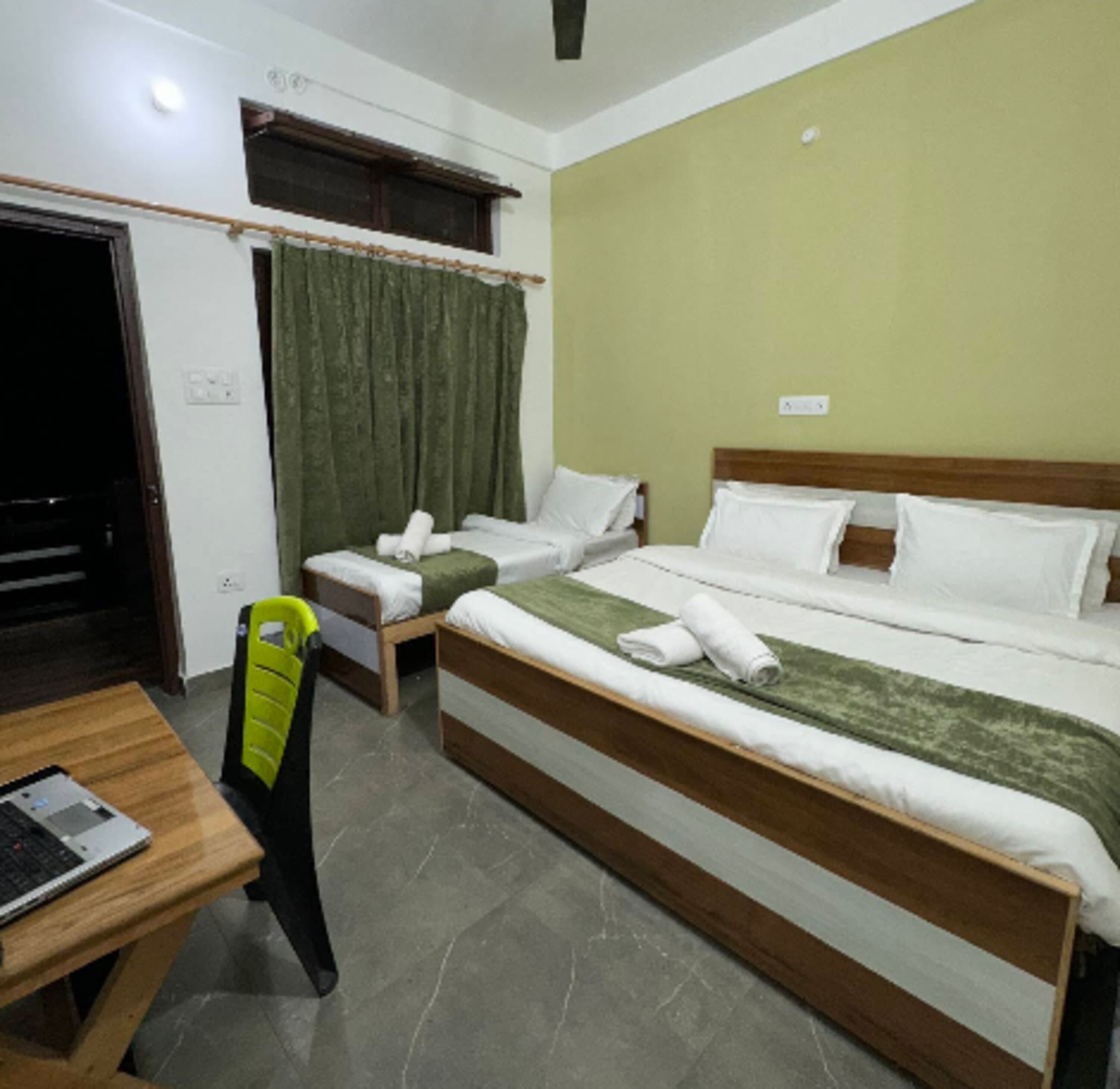 A hotel room with a large bed, two smaller beds, a desk with a laptop, and a chair.
