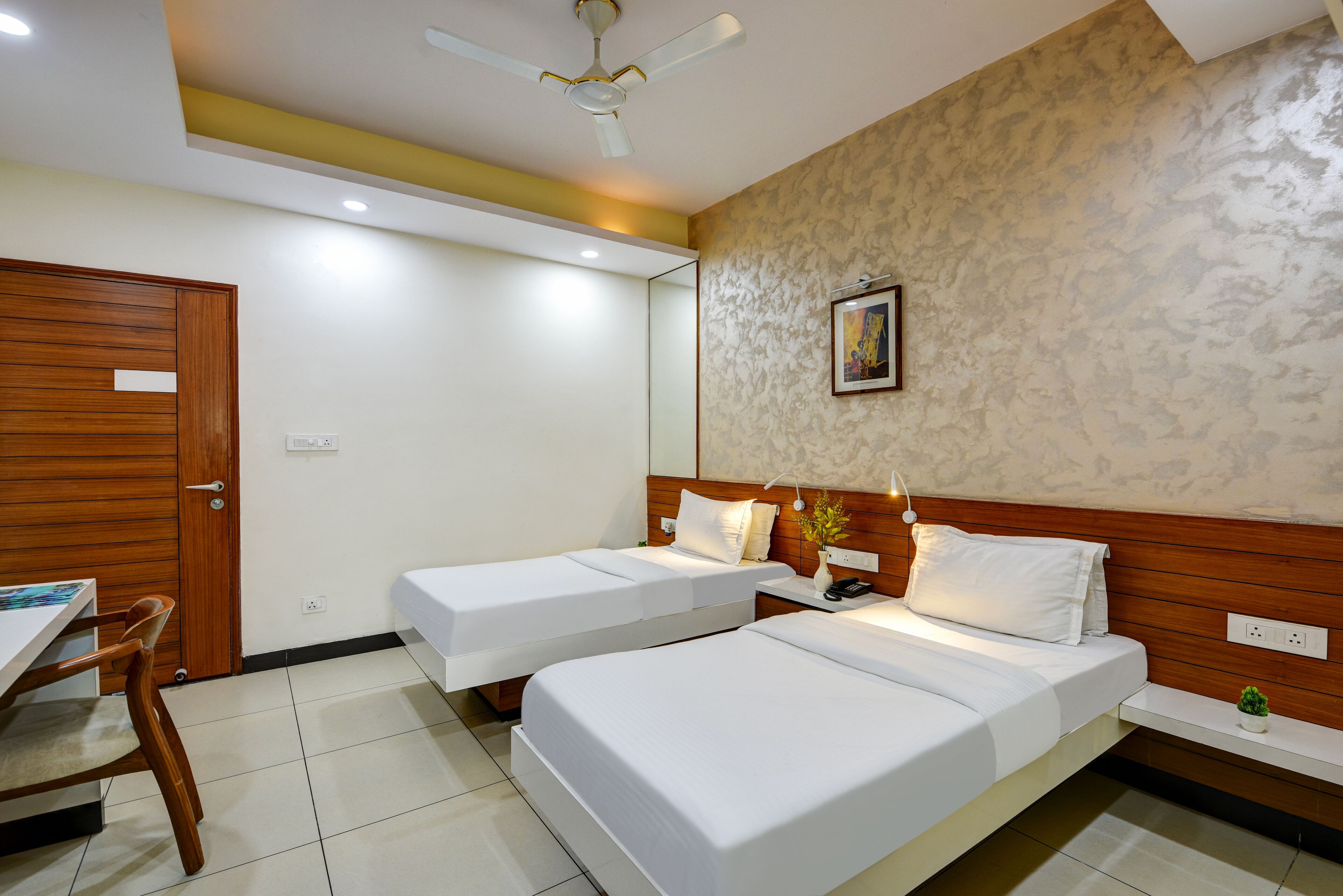 A hotel room with two single beds, a wooden desk, a chair, and a ceiling fan.