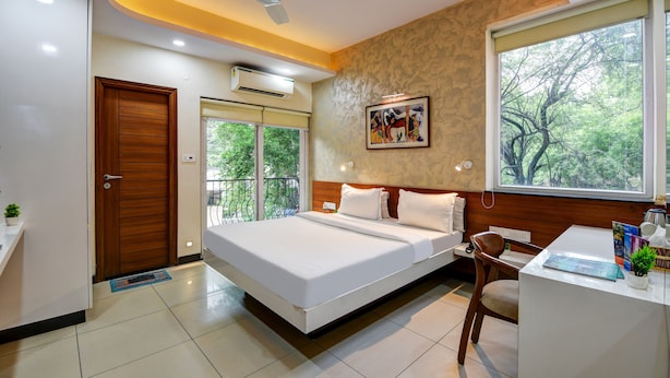 Una habitación de hotel moderna con cama, escritorio, silla, ventilador de techo y una ventana grande con vista a árboles.