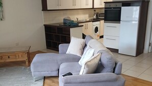 Una sala moderna con un sofá seccional gris, una mesa de centro de madera y una alfombra gris sobre piso de madera. Se observa una cocina con gabinetes y electrodomésticos blancos al fondo.