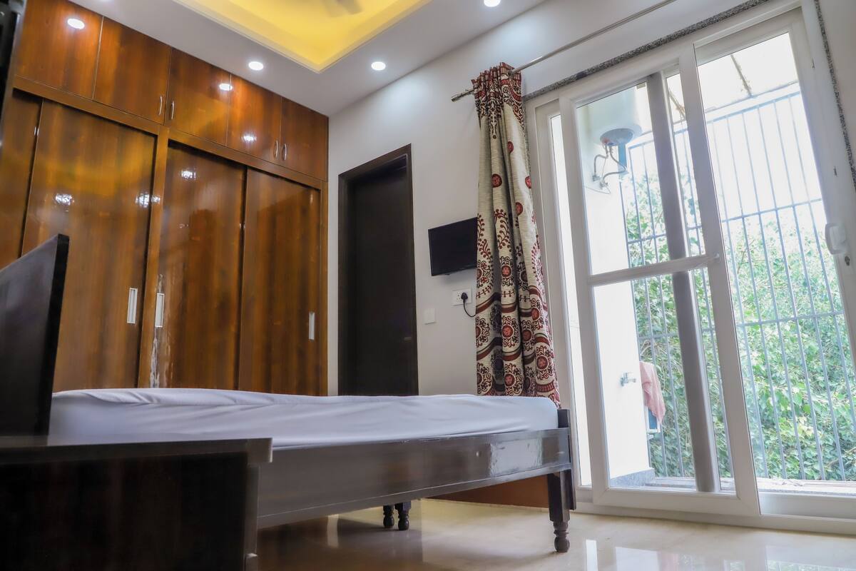 A bedroom with a bed, wooden wardrobe, a television, and a large window with a view of greenery.
