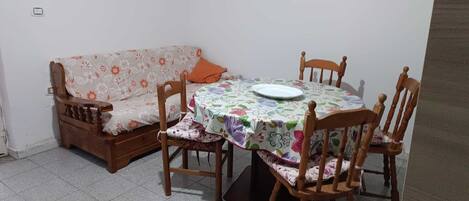 A dining area with a round table, floral tablecloth, wooden chairs, and a sofa with a floral pattern.