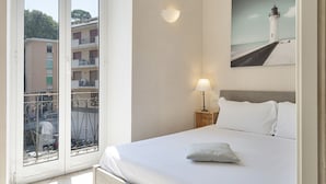 Hotelkamer met een bed, nachtkastje met een lamp, schilderij van een vuurtoren en een balkon met uitzicht op gebouwen en een boom.