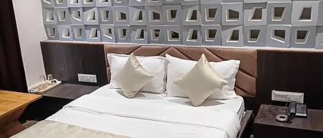 A modern hotel room with a large bed, a headboard with geometric cutouts, a bedside table with a phone, and a nightstand with a lamp.
