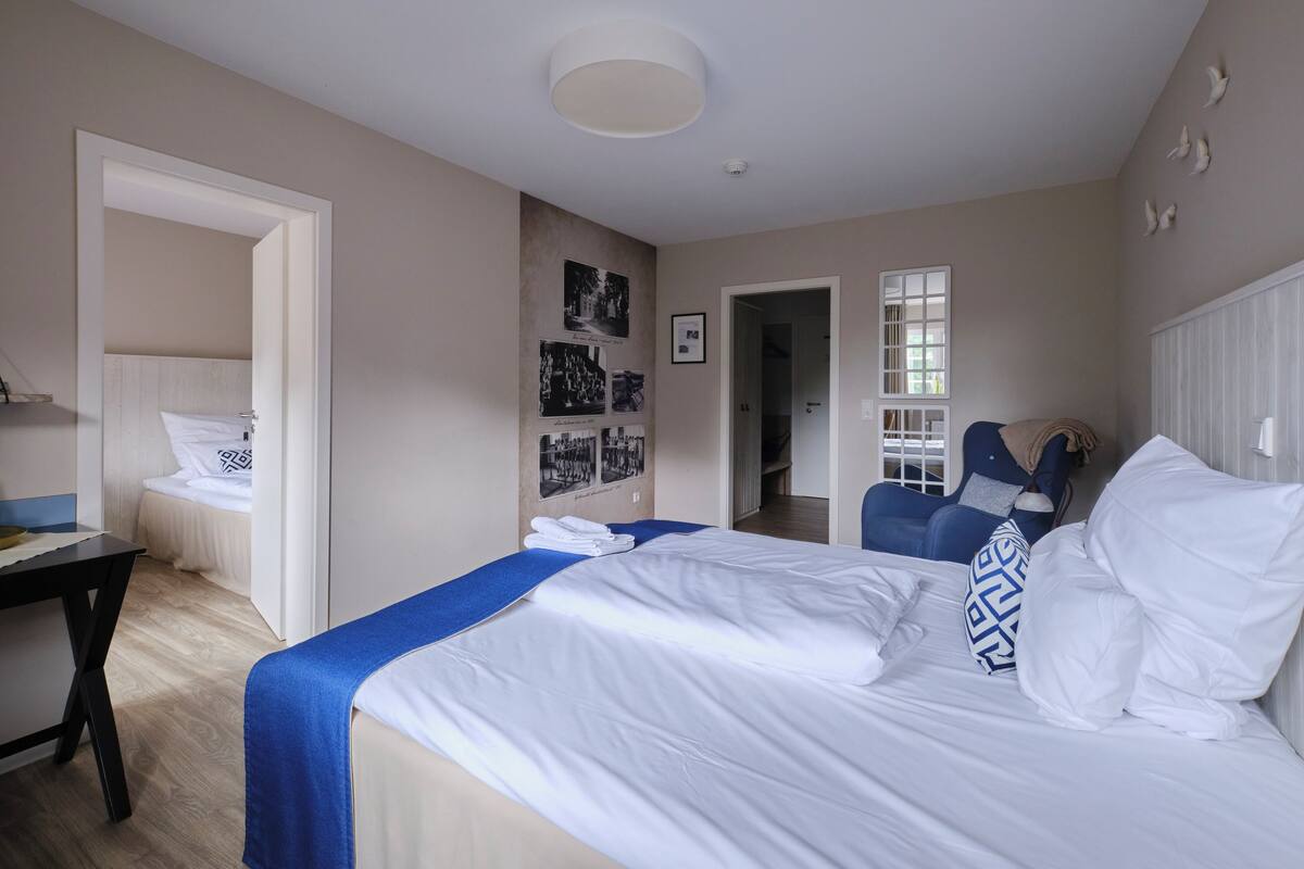Une chambre d’hôtel avec un lit, un bureau et une chaise. Il y a des tableaux au mur et une fenêtre avec des stores.