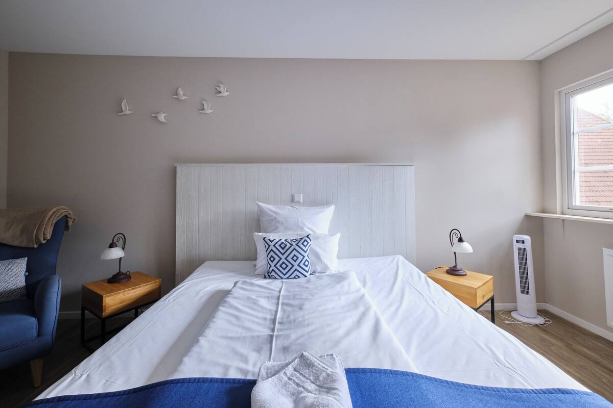 Un lit bien fait, avec du linge de lit blanc, un plaid bleu et une serviette blanche. On y trouve une table de chevet en bois avec une lampe, une chaise bleue et un radiateur blanc.