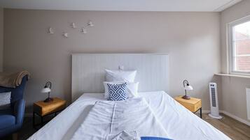 A neatly made bed with white bedding, a blue throw blanket, and a white towel. There is a wooden nightstand with a lamp, a blue chair, and a white radiator.