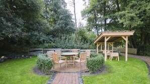 A patio with a wooden table and chairs surrounded by a brick pathway, a wooden pergola, and a wooden fence in a garden setting. - Hotel Utspann - Schafflund (Schafflund)