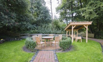 Eine Terrasse mit Holztisch und -stühlen, umgeben von einem Ziegelweg, einer Holzpergola und einem Holzzaun in einem Garten.