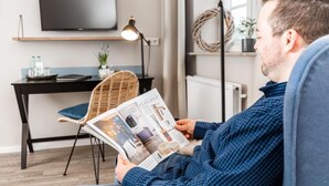A person reading a magazine in a room with a modern design, featuring a black desk, wicker chair, and a flat-screen TV on the wall. - Hotel Utspann - Schafflund (Schafflund)