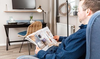 Eine Person liest eine Zeitschrift in einem Zimmer mit modernem Design, das einen schwarzen Schreibtisch, einen Korbstuhl und einen Flachbildfernseher an der Wand zeigt.