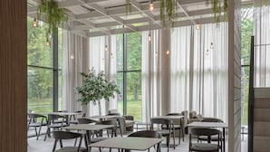 Un espacio interior amplio con mesas y sillas dispuestas para comer, ventanas grandes que ofrecen vistas a la vegetación y plantas colgantes que aportan un toque natural.