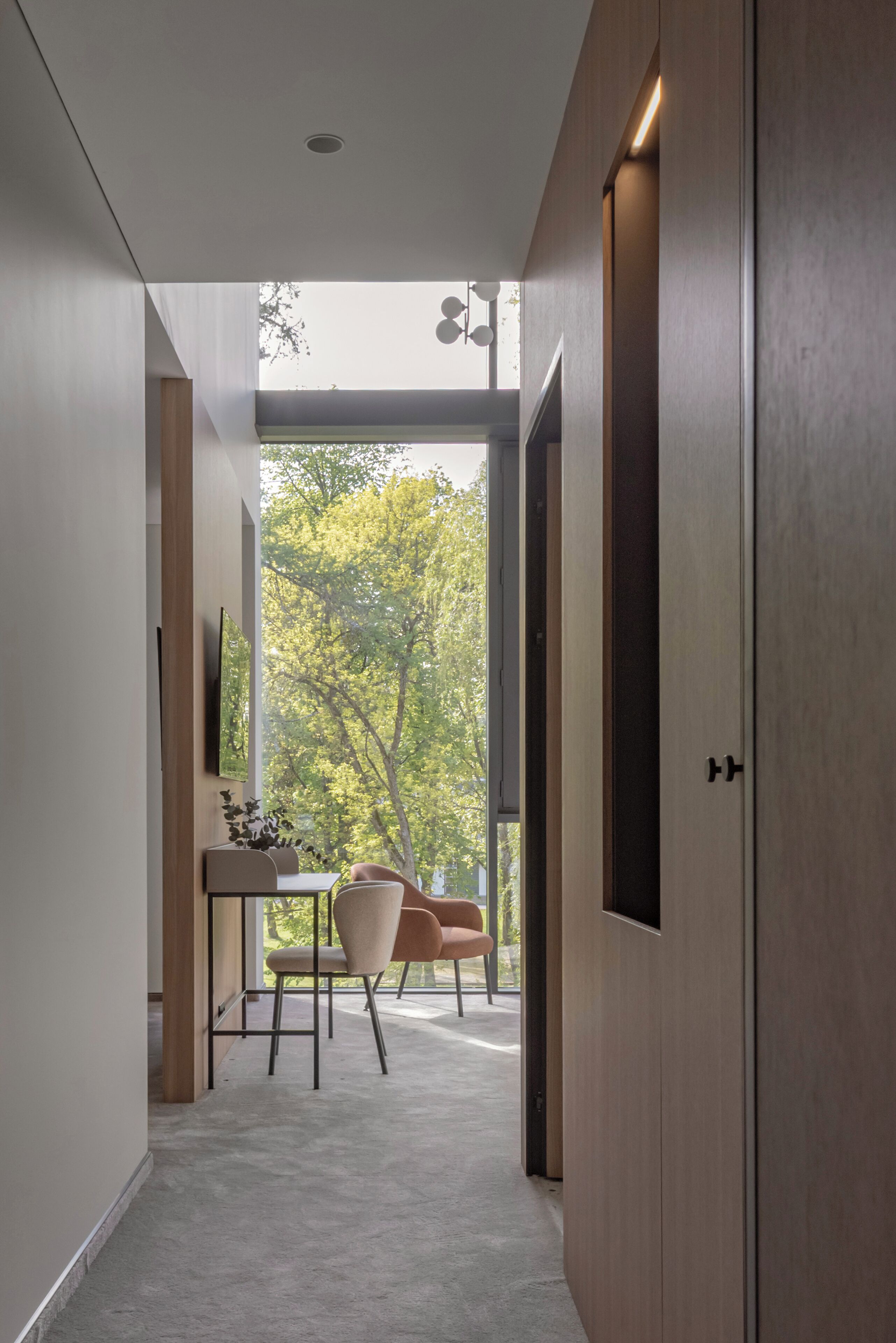 A modern hallway with a view of trees through a large window, a desk with a chair, and a door.
