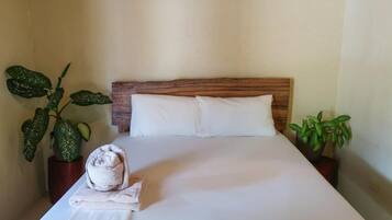A neatly made bed with white linens, two pillows, and a rolled towel on a wooden headboard. There are two potted plants, one on each side of the bed.