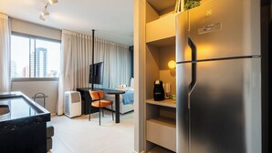 Una habitación de hotel moderna con un refrigerador grande, un escritorio con una silla, una cama, un televisor y una ventana con cortinas.