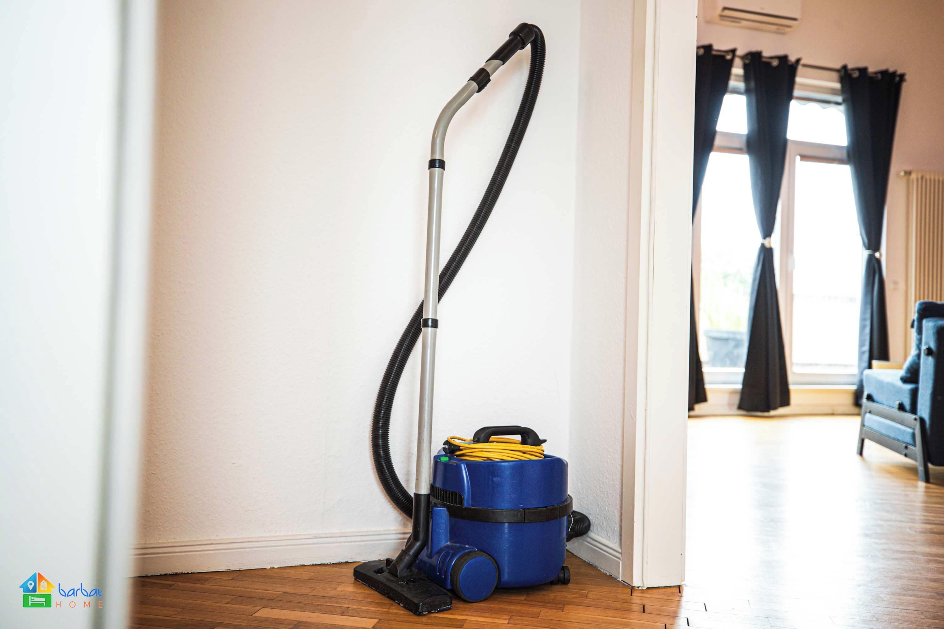A vacuum cleaner in a hallway with a wooden floor, a window with curtains, and a radiator.