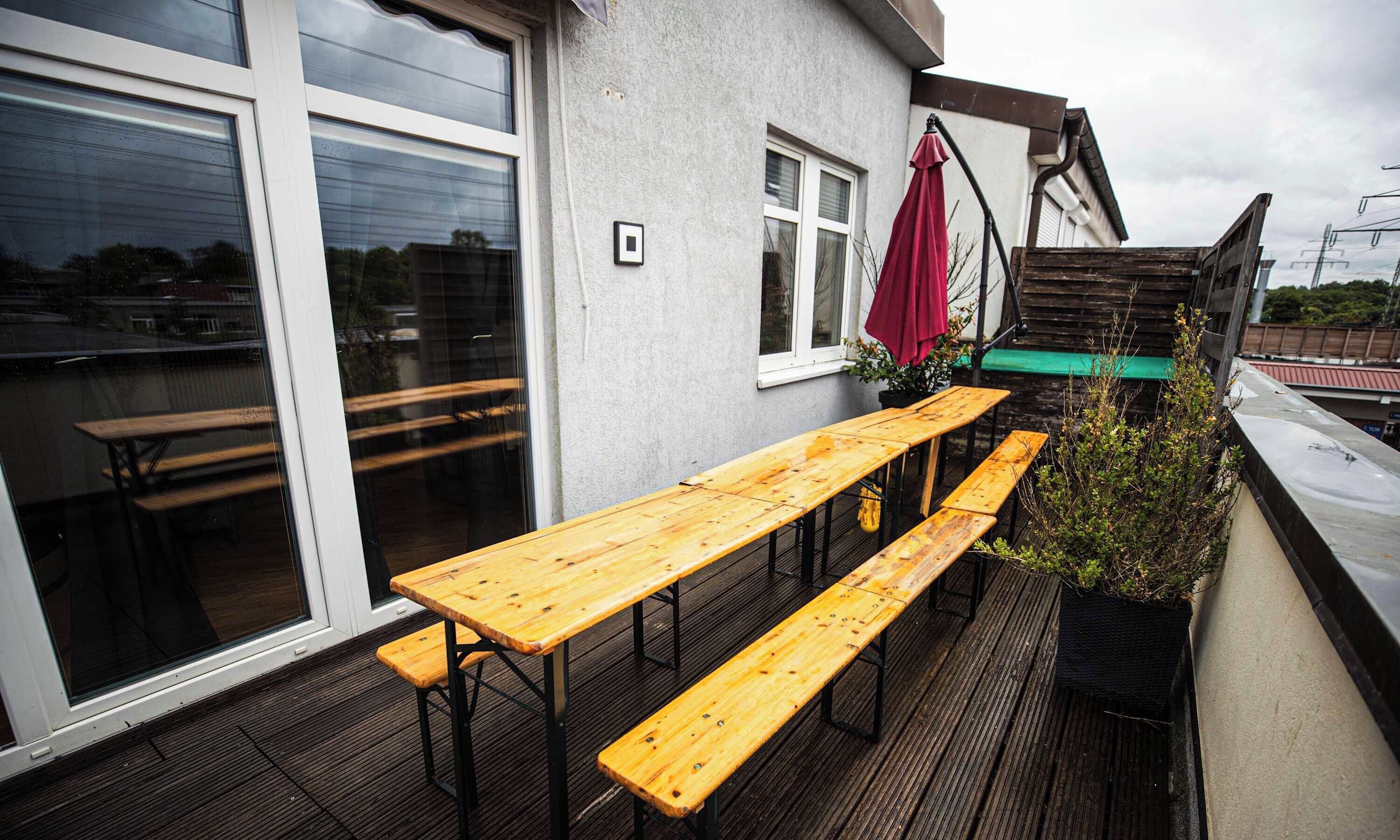 Eine Terrasse mit Holztischen und -bänken, einem roten Sonnenschirm und einem Blumentopf.