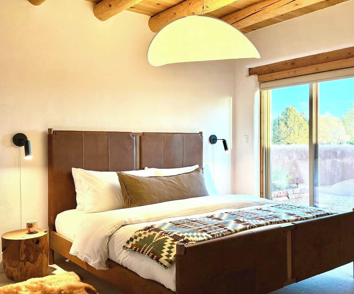 Ein Schlafzimmer mit einem großen Bett, einem Holzkopfteil, Nachttischlampen und Blick ins Freie durch ein großes Fenster.