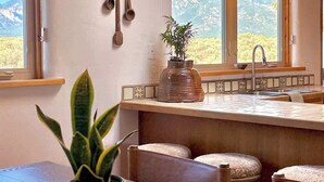 A rustic kitchen with wooden beams, a large window with a mountain view, a hanging light fixture, and a plant on the counter.