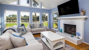 A living room with a white fireplace, a flat-screen TV, a beige sofa with patterned cushions, and large windows offering a view of a golf course.