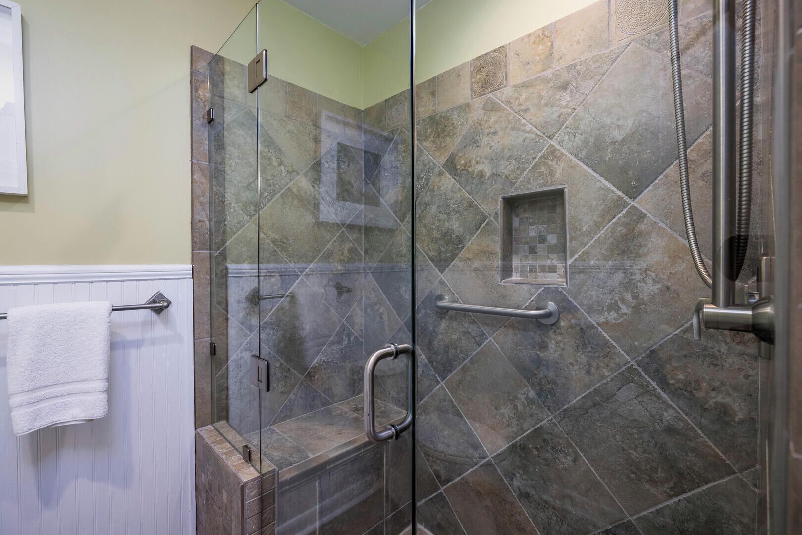 Um chuveiro com paredes de vidro, corrimão, porta-toalhas e um pequeno detalhe em azulejos quadrados na parede.
