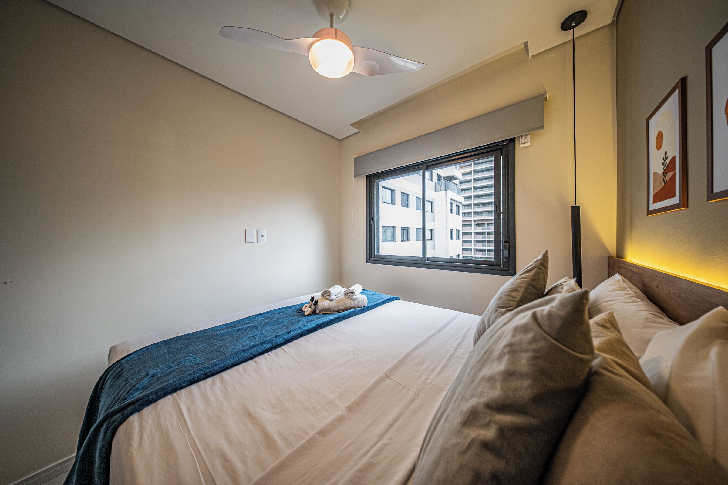 A bedroom with a bed, a ceiling fan, a window with blinds, and a wall-mounted light fixture.