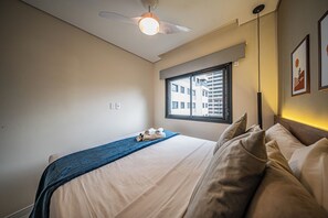 Une chambre à coucher comprenant un lit, un ventilateur de plafond, une fenêtre avec des stores et un luminaire fixé au mur.