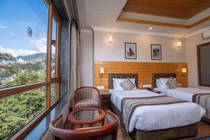 Habitación de hotel con dos camas, cabecera de madera, una mesa redonda y vistas a un paisaje urbano a través de grandes ventanales.