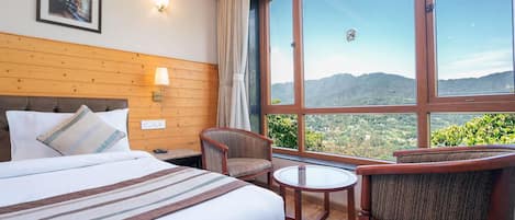 A hotel room with a large bed, two chairs, a small table, and a large window offering a mountain view.