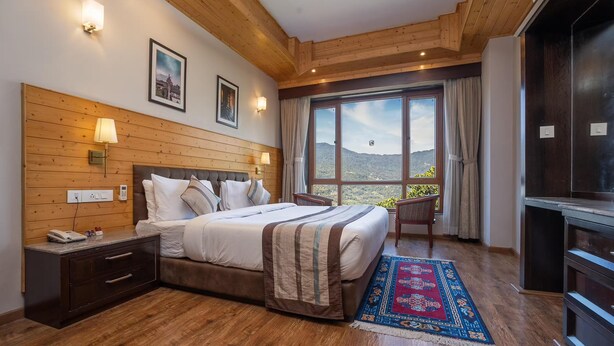 A bedroom with a wooden headboard, a bed with a striped blanket, a nightstand with a phone, a desk with a lamp, a large window with a view of mountains, and a dark wooden cabinet. - Voyage Alpine Abode (Gangtok)