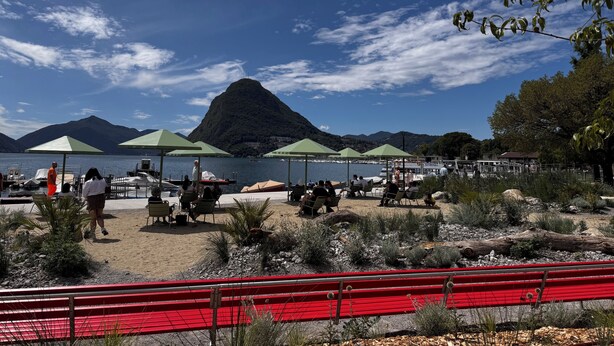A lakeside beach with green umbrellas, a red bench, and mountains in the background. - Lugano Lakeside: The Nest - your refuge in Castagnola (Lugano)